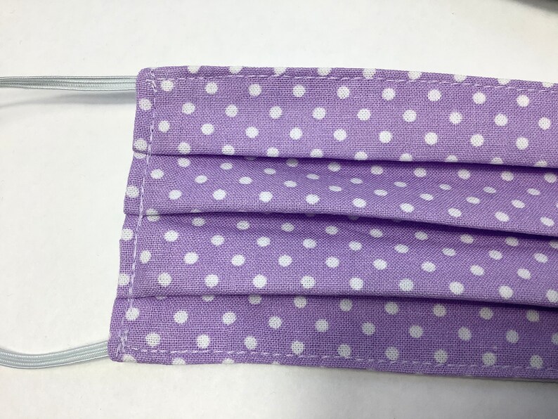 May include: A purple face mask with white polka dots. The mask is pleated and has elastic ear loops.