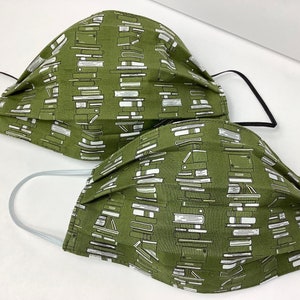 May include: Two green fabric face masks with a white print of stacked books. The masks have elastic ear loops.