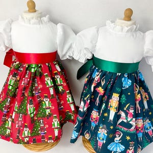 May include: Two white doll dresses with puffed sleeves and a ruffled collar. The dresses have a red and green Nutcracker print skirt with a satin ribbon belt.