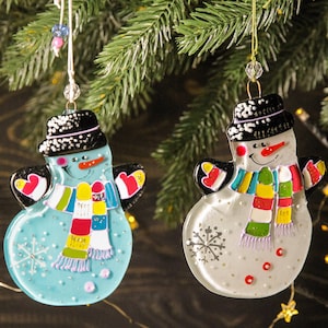 May include: Two glass snowman ornaments with black hats, colorful scarves, and snowflake designs. One snowman is blue and the other is white.