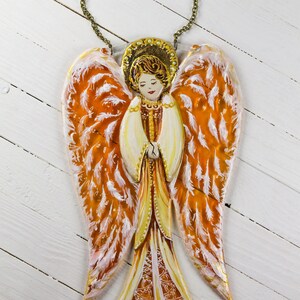 May include: A hand-painted wooden angel ornament with orange and white wings. The angel is wearing a gold dress and has a halo. The ornament is hanging from a gold chain.