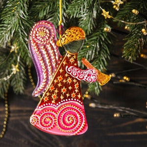 May include: A glass angel ornament with a pink, red, and gold painted design. The angel is playing a trumpet and has a halo.