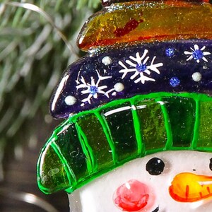 Puede incluir: Un adorno de vidrio de mu&ntilde;eco de nieve con un sombrero verde, un sombrero a rayas morado y naranja y copos de nieve blancos. El mu&ntilde;eco de nieve tiene una cara blanca con ojos negros, una nariz roja y una nariz de zanahoria naranja.