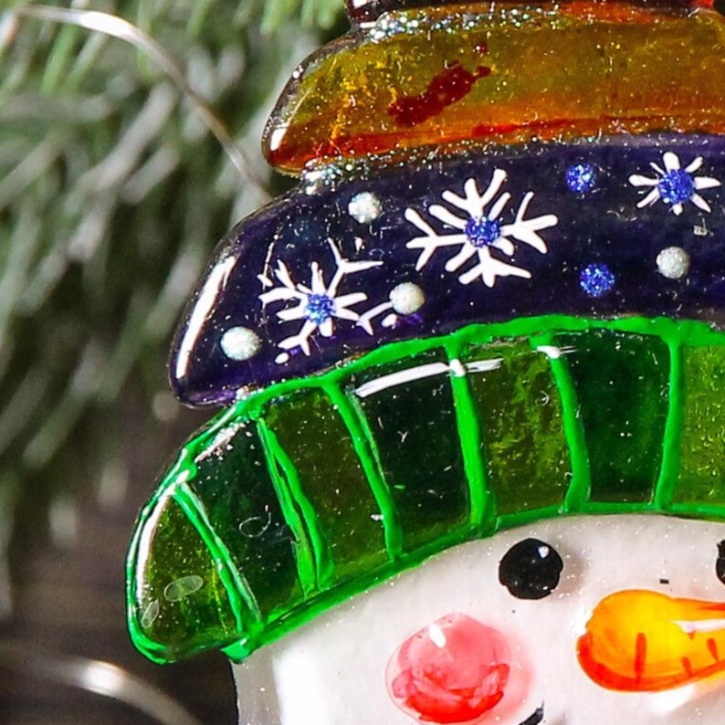Puede incluir: Un adorno de vidrio de mu&ntilde;eco de nieve con un sombrero verde, un sombrero a rayas morado y naranja y copos de nieve blancos. El mu&ntilde;eco de nieve tiene una cara blanca con ojos negros, una nariz roja y una nariz de zanahoria naranja.