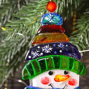 Puede incluir: Un adorno de vidrio de mu&ntilde;eco de nieve con un sombrero verde, una bufanda azul y una cara blanca. El adorno est&aacute; decorado con cuentas de colores y copos de nieve.