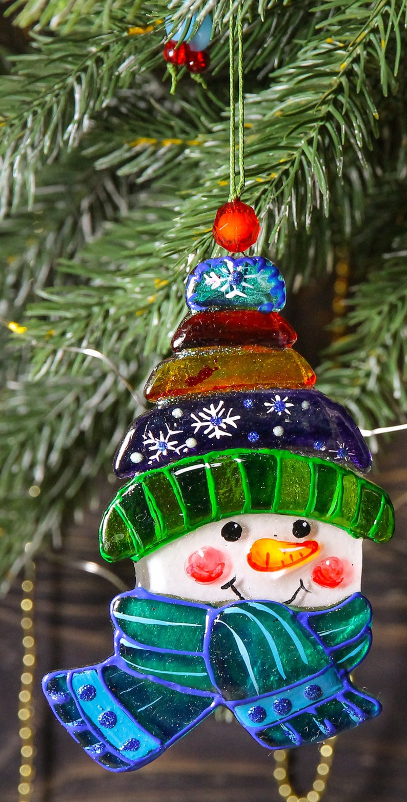 Puede incluir: Un adorno de vidrio de mu&ntilde;eco de nieve con un sombrero verde, una bufanda azul y una cara blanca. El adorno est&aacute; decorado con cuentas de colores y copos de nieve.