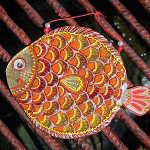May include: A colorful, hand-painted fish ornament with red, orange, yellow, and gold scales. The fish is decorated with white beads and has a red string attached to it.
