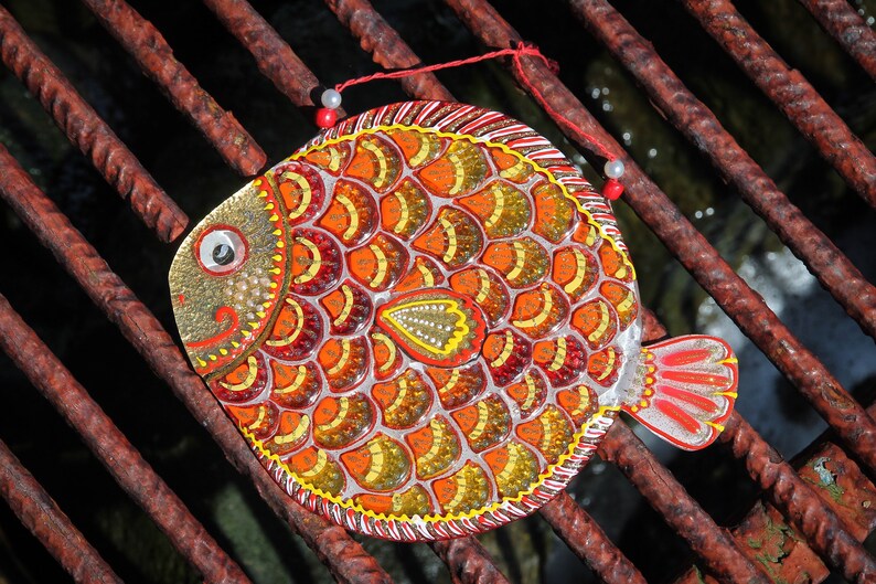 May include: A colorful, hand-painted fish ornament with red, orange, yellow, and gold scales. The fish is decorated with white beads and has a red string attached to it.