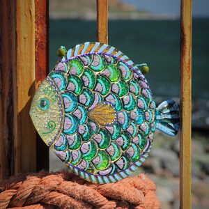 May include: A colorful, hand-painted fish ornament with green, blue, and purple scales. The fish is made of glass and has a shiny, iridescent finish. It is hanging from a rusty metal gate.