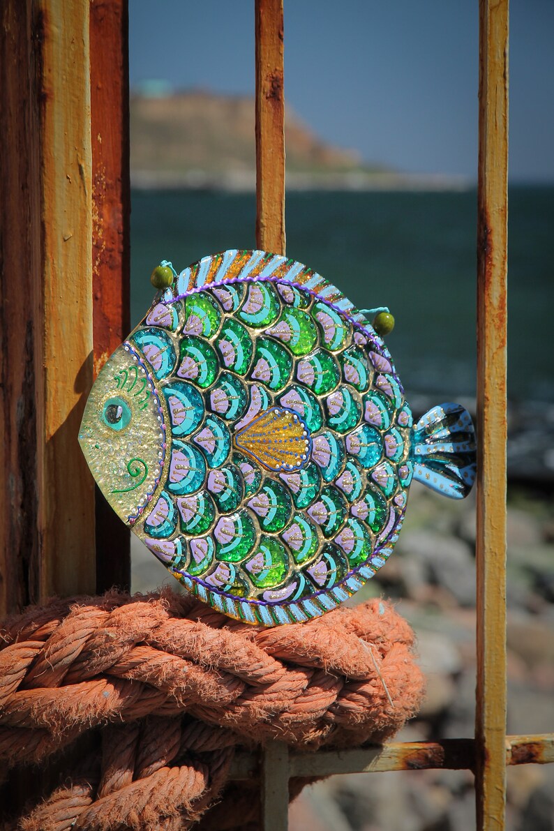 May include: A colorful, hand-painted fish ornament with green, blue, and purple scales. The fish is made of glass and has a shiny, iridescent finish. It is hanging from a rusty metal gate.