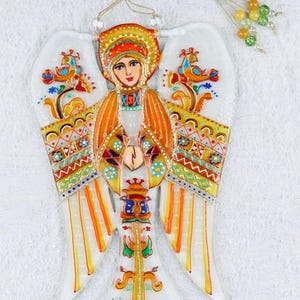 May include: A colorful glass angel ornament with a painted image of a woman in a traditional headdress. The angel has orange, yellow, and white wings with intricate patterns. The ornament has a string for hanging.