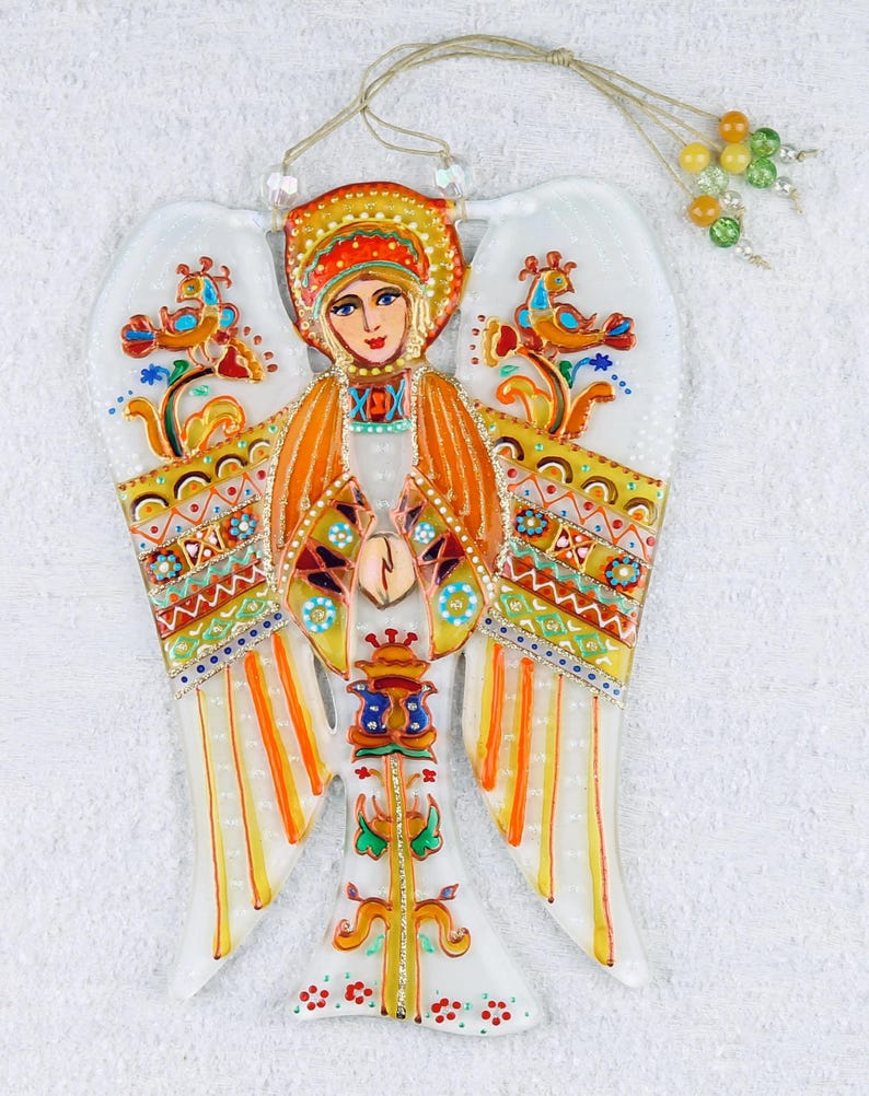 May include: A colorful glass angel ornament with a painted image of a woman in a traditional headdress. The angel has orange, yellow, and white wings with intricate patterns. The ornament has a string for hanging.