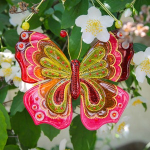 Puede incluir: Un adorno de mariposa de vidrio colorido con acentos rojos, amarillos, verdes y marrones. La mariposa est&aacute; colgada de una rama de un arbusto de flores blancas.