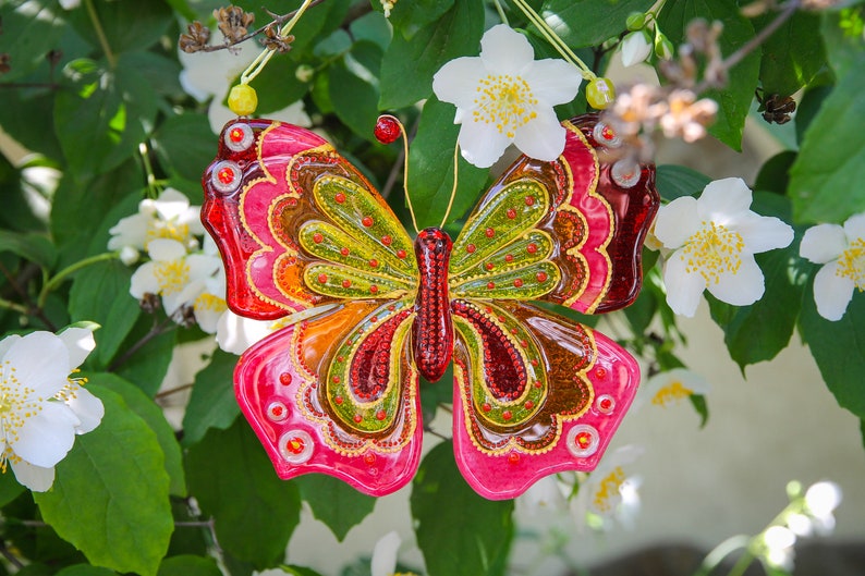 Puede incluir: Un adorno de mariposa de vidrio colorido con acentos rojos, amarillos, verdes y marrones. La mariposa est&aacute; colgada de una rama de un arbusto de flores blancas.