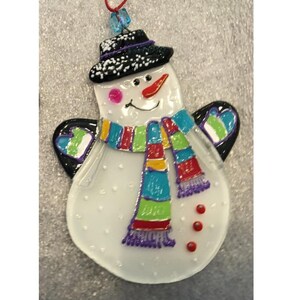 Pode incluir: Enfeite de boneco de neve com cartola preta, cachecol colorido e nariz de cenoura. O boneco de neve tem asas pretas com detalhes verdes e azuis. O enfeite &eacute; branco com detalhes vermelhos, rosa e roxos. Uma corda vermelha est&aacute; presa para pendurar.