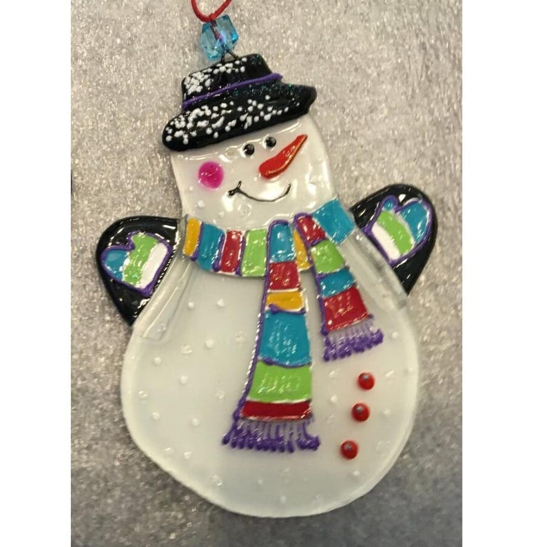 Pode incluir: Enfeite de boneco de neve com cartola preta, cachecol colorido e nariz de cenoura. O boneco de neve tem asas pretas com detalhes verdes e azuis. O enfeite &eacute; branco com detalhes vermelhos, rosa e roxos. Uma corda vermelha est&aacute; presa para pendurar.