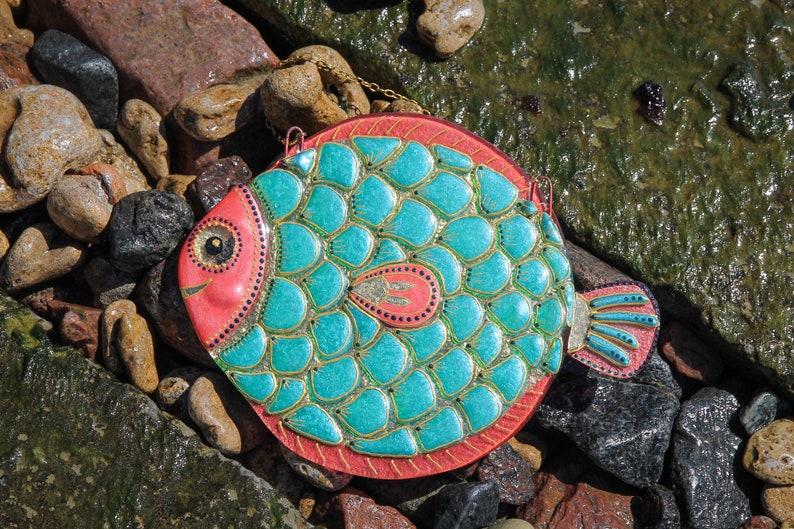 May include: A turquoise and pink fish-shaped wall hanging with gold accents. The fish has a scaly design and a large eye.