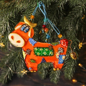 May include: A colorful patchwork cow ornament with a red, orange, yellow, green, blue, and white design. The ornament is hanging from a blue and white string with beads.