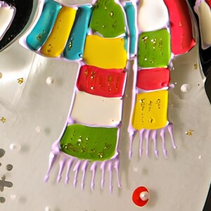 May include: A close-up of a colorful striped scarf with a purple fringe. The scarf is made of painted glass and has a white, red, green, yellow, and blue design.