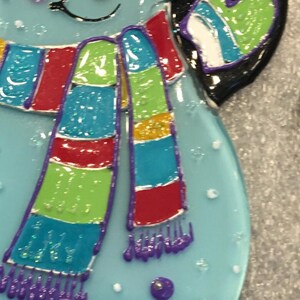 May include: A blue glass snowman ornament with a colorful striped scarf. The scarf has red, yellow, green, blue, and purple stripes. The snowman has a white face with black eyes and a black mouth.