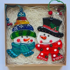 Puede incluir: Dos adornos de vidrio de mu&ntilde;eco de nieve en una caja con papel triturado. Un mu&ntilde;eco de nieve lleva un gorro y una bufanda azules y verdes, el otro lleva un gorro y una bufanda rojos y blancos.