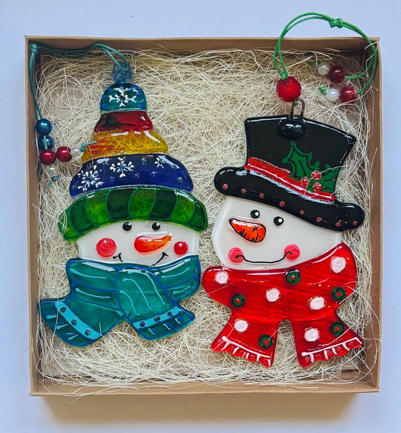 Puede incluir: Dos adornos de vidrio de mu&ntilde;eco de nieve en una caja con papel triturado. Un mu&ntilde;eco de nieve lleva un gorro y una bufanda azules y verdes, el otro lleva un gorro y una bufanda rojos y blancos.