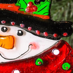 Puede incluir: Un adorno de vidrio de mu&ntilde;eco de nieve con un sombrero negro, acebo verde y bayas rojas. El mu&ntilde;eco de nieve tiene una cara blanca con ojos negros, una nariz naranja y un rubor rojo.