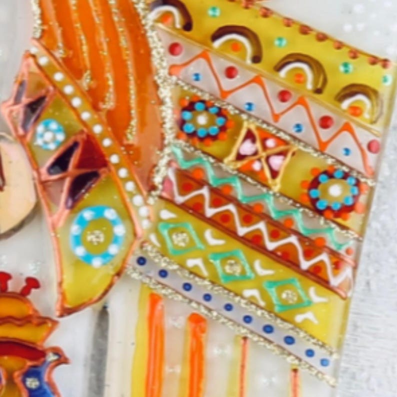 May include: A close-up of a colorful, hand-painted glass ornament with a geometric pattern. The ornament features a variety of colors, including yellow, orange, red, green, blue, and white. The ornament is decorated with small, white dots and a gold glitter finish.