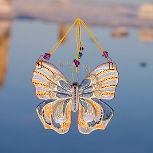 Puede incluir: Un adorno de mariposa decorativo con detalles dorados y plateados, colgado de una cuerda con cuentas moradas y azules.