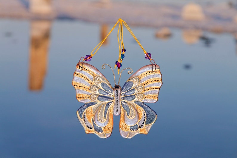 Puede incluir: Un adorno de mariposa decorativo con detalles dorados y plateados, colgado de una cuerda con cuentas moradas y azules.