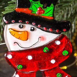 Puede incluir: Un adorno de vidrio de mu&ntilde;eco de nieve con un sombrero de copa negro, una bufanda de lunares rojos y blancos y detalles de acebo verde.