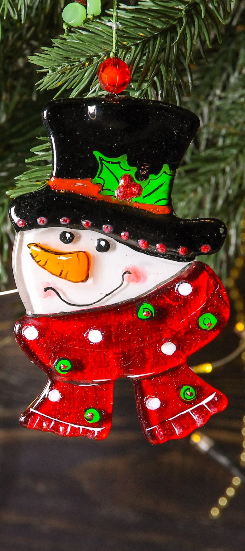 Puede incluir: Un adorno de vidrio de mu&ntilde;eco de nieve con un sombrero de copa negro, una bufanda de lunares rojos y blancos y detalles de acebo verde.
