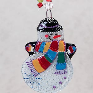 Pode incluir: Enfeite de Natal de boneco de neve com chap&eacute;u preto e cachecol colorido. O boneco de neve tem um nariz vermelho e um cachecol com listras azuis, verdes, amarelas e vermelhas. O enfeite &eacute; decorado com pontos brancos e detalhes roxos.