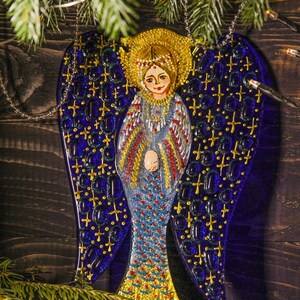 May include: A blue glass angel ornament with gold accents and a painted face. The angel has a halo and is wearing a white dress with gold trim. The ornament is hanging from a silver chain.