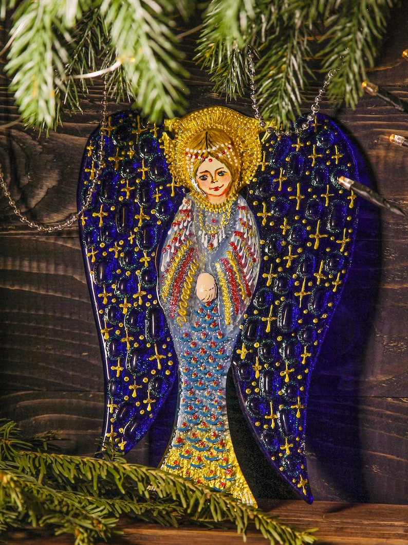 May include: A blue glass angel ornament with gold accents and a painted face. The angel has a halo and is wearing a white dress with gold trim. The ornament is hanging from a silver chain.