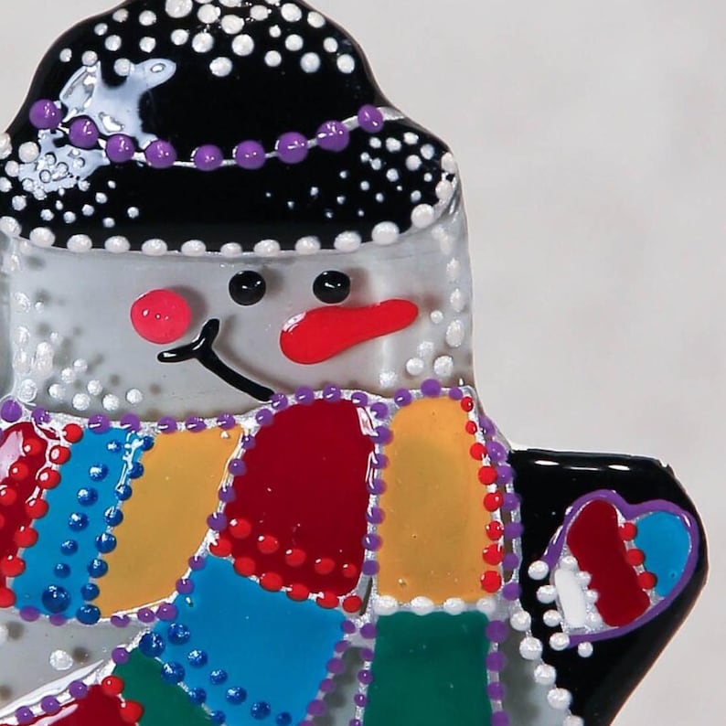 Pode incluir: Um enfeite de boneco de neve extravagante com um chap&eacute;u preto adornado com bolinhas brancas e contas roxas. O boneco de neve tem um nariz vermelho e um cachecol colorido. O enfeite tem um desenho em forma de cora&ccedil;&atilde;o na lateral.