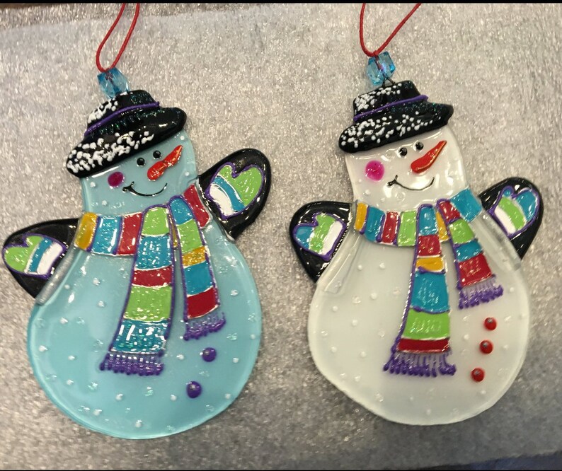 May include: Two glass snowman ornaments with black hats, white faces, and colorful scarves. The snowman on the left is blue and the snowman on the right is white.