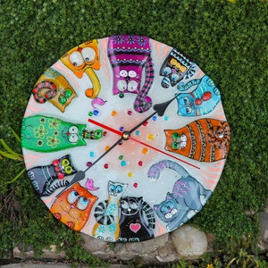 May include: A white round clock with colorful painted cats around the face. The clock hands are black and red. The background is a white with colorful dots.