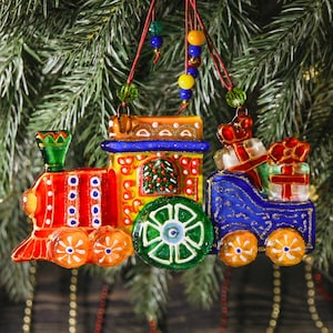 May include: A colorful glass Christmas ornament shaped like a train. The train is red, yellow, and blue with orange wheels and is decorated with gifts and a Christmas tree. The ornament hangs from a red string with colorful beads.