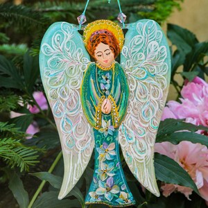 May include: A teal blue angel ornament with white and gold painted wings and a floral design. The angel is holding its hands together in prayer. The ornament is hanging from a string with beads.