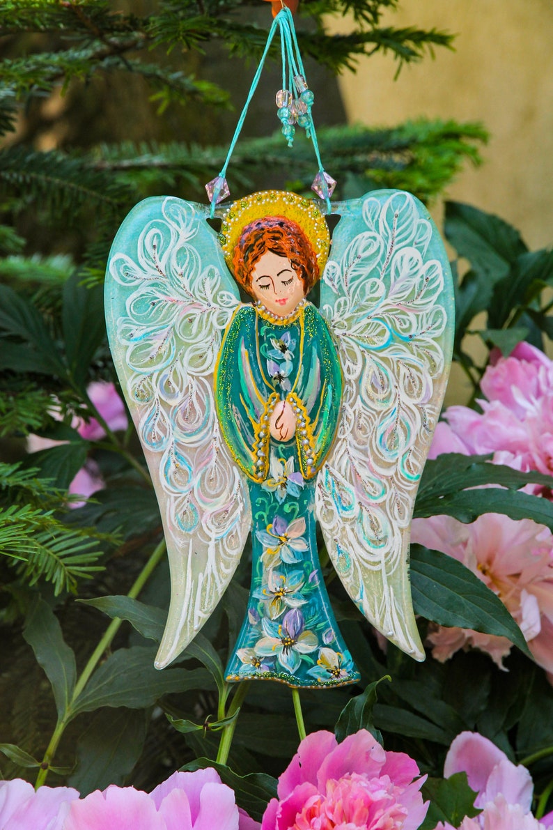 May include: A teal blue angel ornament with white and gold painted wings and a floral design. The angel is holding its hands together in prayer. The ornament is hanging from a string with beads.