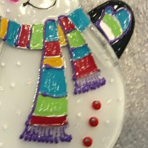 May include: A white snowman ornament with a colorful striped scarf and purple fringe. The snowman has three red buttons and a black hat.