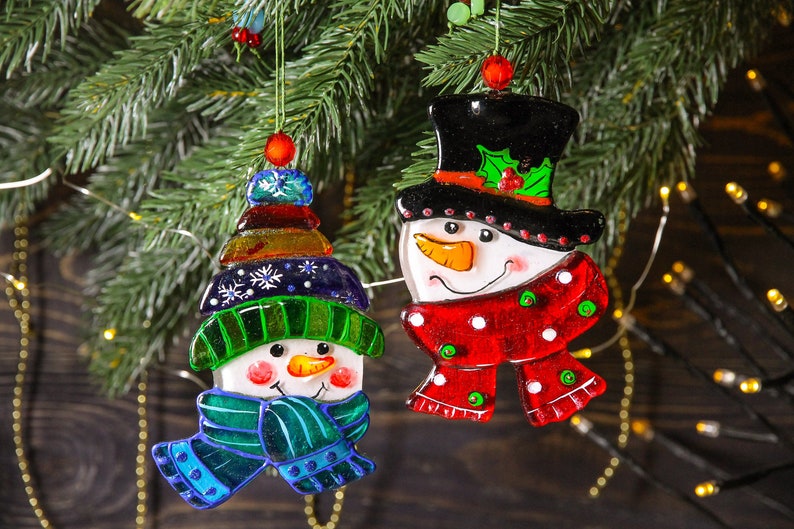 Puede incluir: Dos adornos de vidrio de mu&ntilde;eco de nieve colgando de un &aacute;rbol de Navidad. Un mu&ntilde;eco de nieve lleva una bufanda roja y blanca y un sombrero de copa negro. El otro mu&ntilde;eco de nieve lleva una bufanda verde y azul y un sombrero azul y verde.