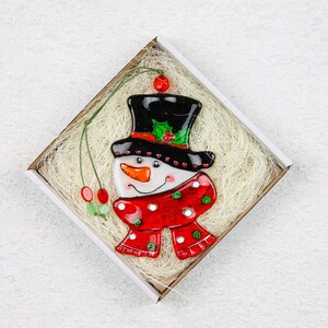Puede incluir: Un adorno de vidrio de mu&ntilde;eco de nieve con un sombrero de copa negro, una bufanda roja y blanca y una cara blanca con una nariz de zanahoria y ojos negros. El adorno est&aacute; en una caja blanca con bordes marrones y material de embalaje blanco.