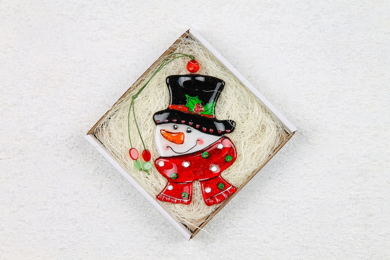 Puede incluir: Un adorno de vidrio de mu&ntilde;eco de nieve con un sombrero de copa negro, una bufanda roja y blanca y una cara blanca con una nariz de zanahoria y ojos negros. El adorno est&aacute; en una caja blanca con bordes marrones y material de embalaje blanco.