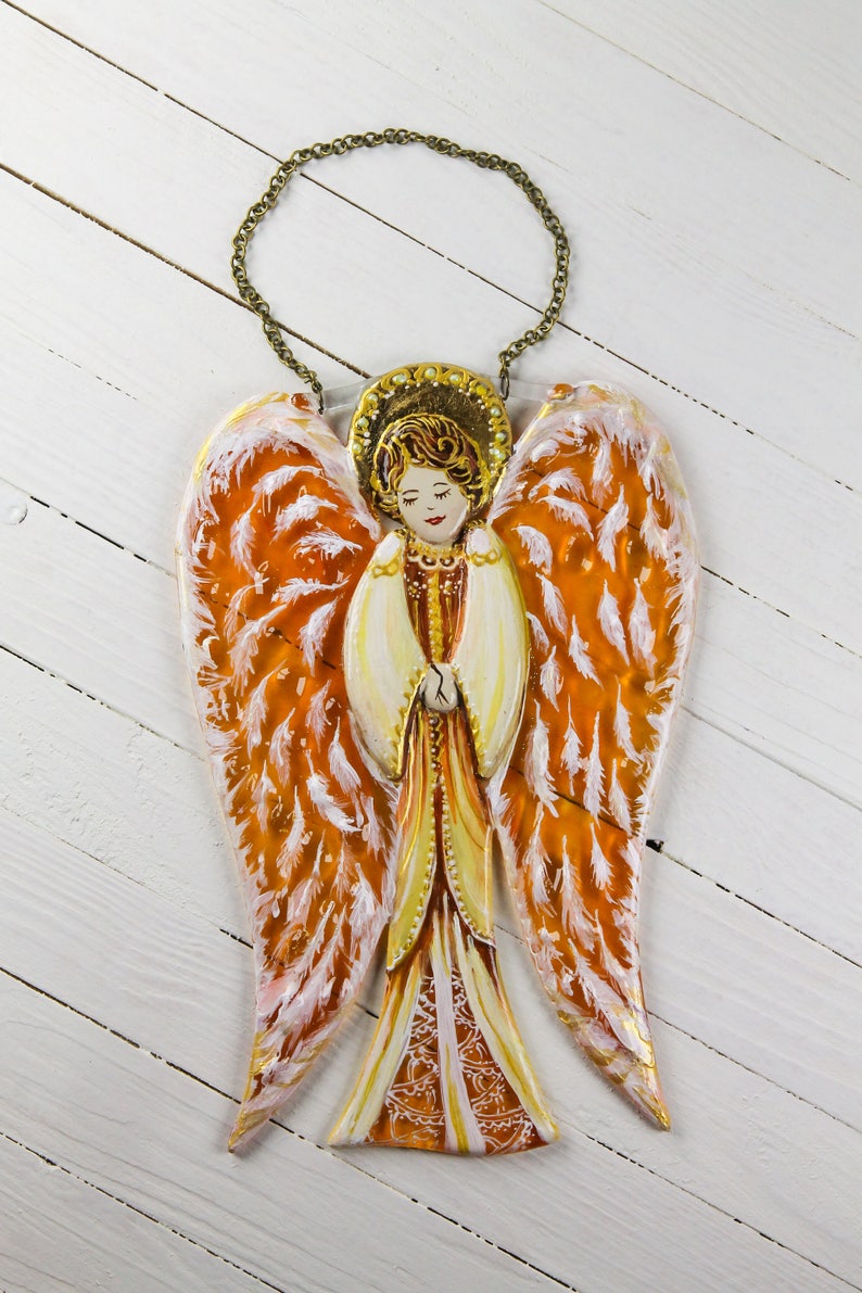 May include: A hand-painted wooden angel ornament with orange and white wings. The angel is wearing a gold dress and has a halo. The ornament is hanging from a gold chain.