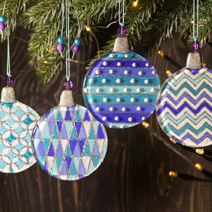 May include: Four glass Christmas ornaments with blue, white, and silver geometric patterns. The ornaments are hanging from a Christmas tree.