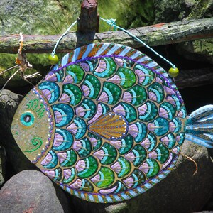 May include: A colorful, hand-painted fish-shaped wall hanging with green, blue, purple, and gold scales. The fish is hanging from a string with green beads.