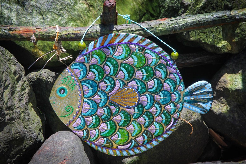 May include: A colorful, hand-painted fish-shaped wall hanging with green, blue, purple, and gold scales. The fish is hanging from a string with green beads.