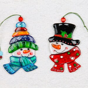 Puede incluir: Dos adornos de vidrio de mu&ntilde;eco de nieve coloridos con detalles verdes, rojos, azules y blancos. El mu&ntilde;eco de nieve de la izquierda tiene un sombrero verde y una bufanda azul. El mu&ntilde;eco de nieve de la derecha tiene un sombrero de copa negro y una bufanda roja.